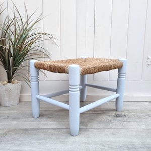 Può includere: Un piccolo sgabello quadrato con un sedile intrecciato marrone chiaro e gambe azzurro pallido. Le gambe sono leggermente tornite con scanalature decorative. Lo sgabello è appoggiato a una parete a pannelli bianchi e un pavimento in legno chiaro.