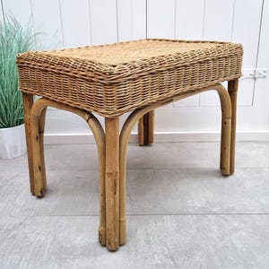 Puede incluir: Un banco rectangular de mimbre con una parte superior tejida y patas de bambú curvadas. El banco es de color marrón claro y es adecuado para uso en interiores. El banco mide aproximadamente 76 cm de largo, 38 cm de ancho y 46 cm de alto.