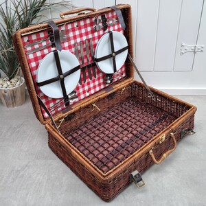 May include: A wicker picnic basket with a red and white checkered lining. The basket is open and contains two white plates, silverware, and a set of wine glasses.