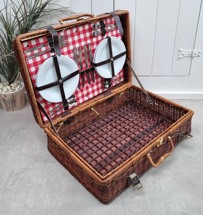 May include: A wicker picnic basket with a red and white checkered lining. The basket is open and contains two white plates, silverware, and a set of wine glasses.