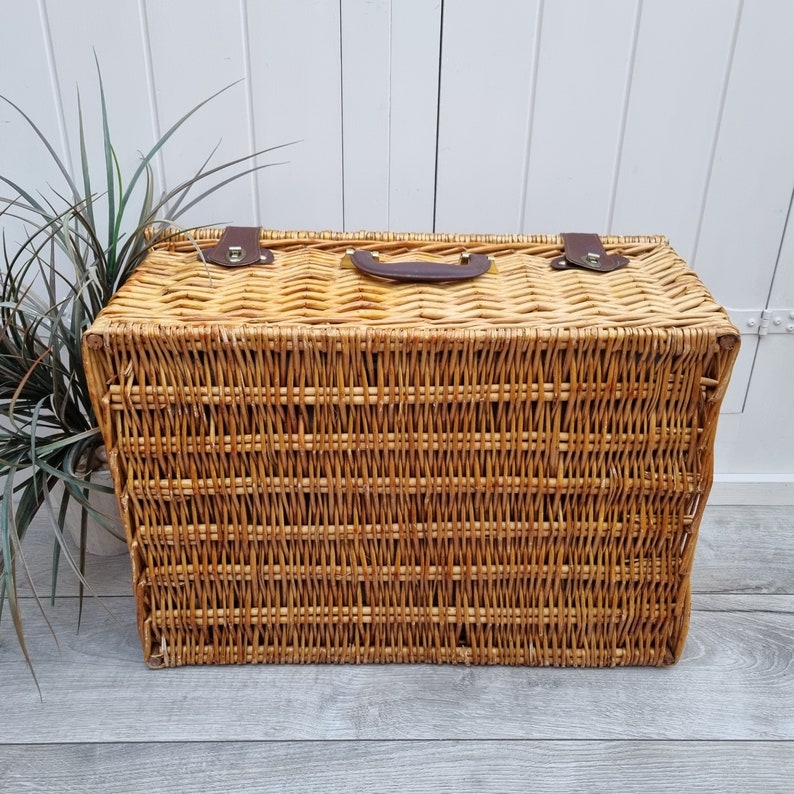 May include: A woven wicker picnic basket with a brown leather handle and metal clasps. The basket is made of natural woven wicker and has a rectangular shape.