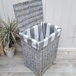 May include: A whitewashed wicker laundry basket with a gray and white striped fabric liner. The lid is open and the basket is sitting on a wood floor.