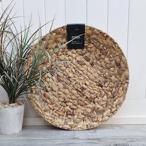 Può includere: Un grande cesto rotondo in paglia di mare intrecciata di colore marrone naturale. Il cesto è fatto di fili di paglia di mare intrecciati strettamente e ha una trama liscia e uniforme. Il cesto è appoggiato su una superficie di legno.