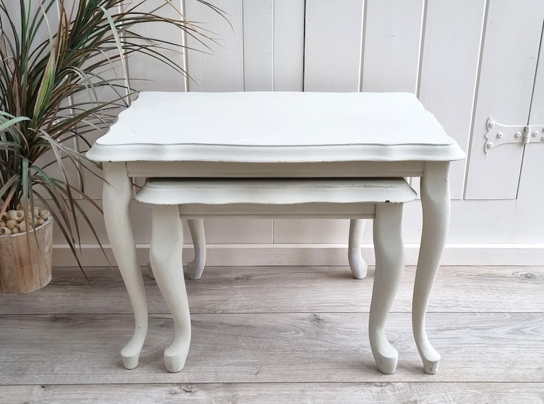 May include: A set of two white nesting tables with a scalloped edge and cabriole legs. The tables are made of wood and have a distressed finish.