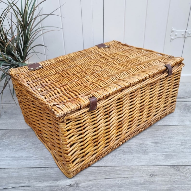 May include: A woven wicker basket with a lid and leather handles. The basket is made of natural brown wicker and has a rectangular shape.