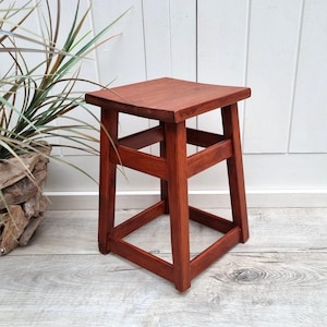 Op de afbeelding: Een kleine, vierkante houten kruk met een roodbruine afwerking. De kruk heeft een eenvoudig, geometrisch ontwerp met een vierkante zitting en vier poten die zijn verbonden door horizontale en schuine steunen. De kruk is ongeveer 30 cm hoog.