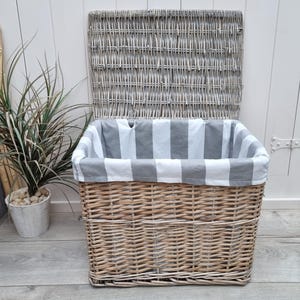 May include: A large, rectangular wicker storage basket with a gray and white striped fabric liner. The lid is open and the basket is sitting on a wooden floor.