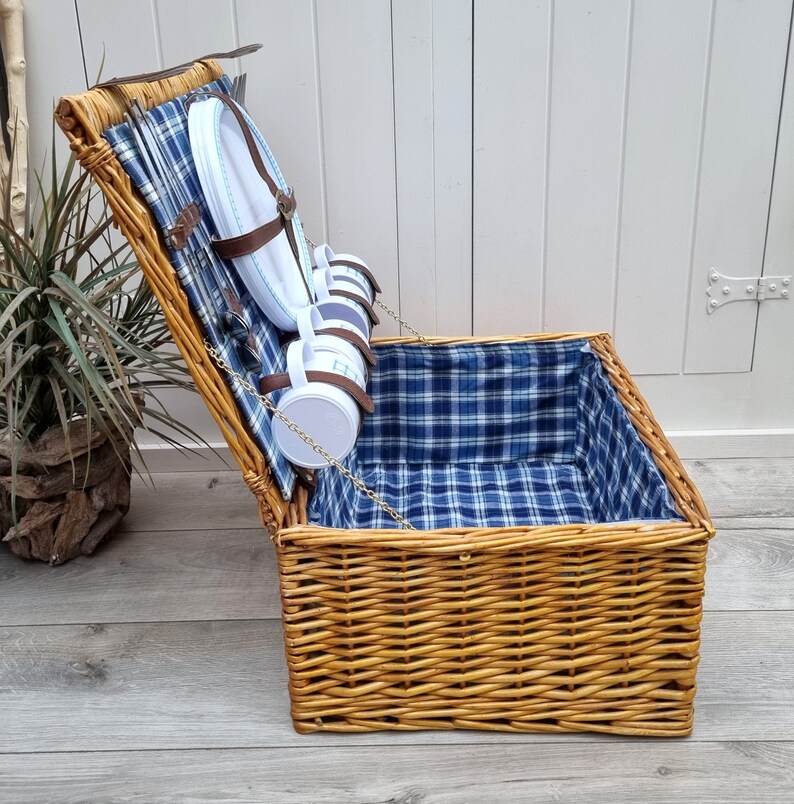 May include: A wicker picnic basket with a blue and white checkered lining. The basket is open and has plates and mugs inside.