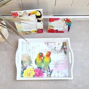 Op de afbeelding: Wit houten dienblad met een bloemen- en vogelontwerp. Het dienblad heeft twee handvatten en heeft een vintage postkaartontwerp met een roze achtergrond en een stempel met de tekst "Post". Het dienblad is versierd met verschillende bloemen, waaronder rozen, lelies en madeliefjes. Er zijn ook twee tortelduifjes op het dienblad, een gele en een groene.