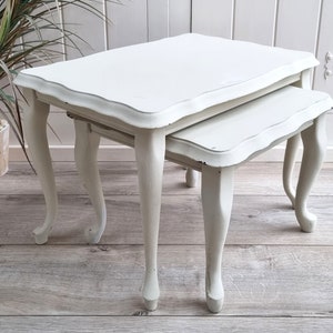 May include: A set of two white nesting tables with a scalloped edge and turned legs. The tables are made of wood and have a distressed finish.
