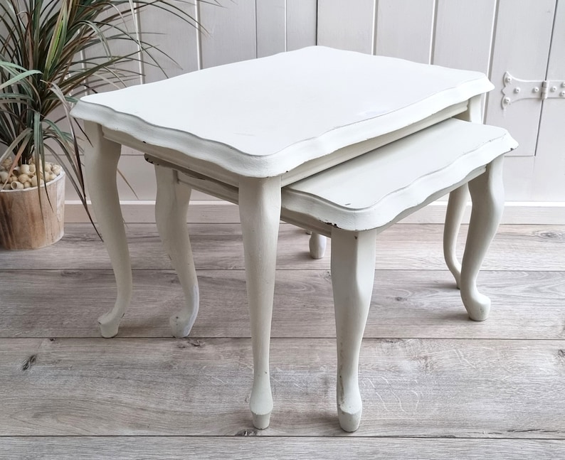 May include: A set of two white nesting tables with a scalloped edge and turned legs. The tables are made of wood and have a distressed finish.