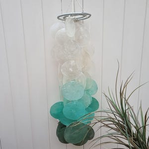 May include: A white and teal seashell wind chime with a wooden ring at the top. The shells are arranged in a gradient from white to teal.