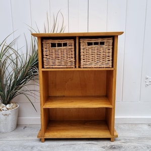 Puede incluir: Una estantería de madera con dos cestas de mimbre en la repisa superior y dos repisas abiertas debajo. La estantería es de color marrón claro y tiene un diseño sencillo.