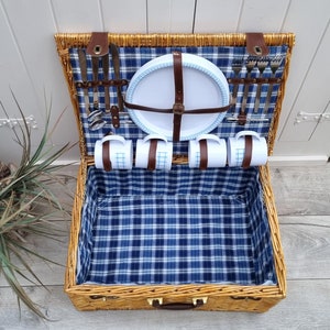 May include: A wicker picnic basket with a blue and white checkered lining. The basket is open and contains a white plate, four mugs, and a set of silverware.