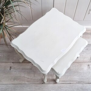 May include: White painted nesting tables with a scalloped edge and turned legs. The tables are stacked on top of each other.