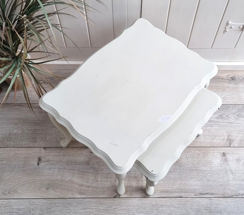 May include: White painted nesting tables with a scalloped edge and turned legs. The tables are stacked on top of each other.