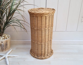 May include: A tall, cylindrical wicker basket with a woven texture and a matching circular lid. The basket features a light tan, natural finish and a sturdy base, constructed from thin, flexible reeds woven in a consistent, repeating pattern.
