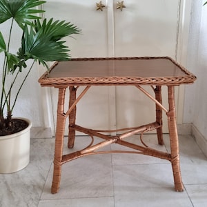 Peut inclure: Une table d'appoint carrée en osier marron avec un plateau en verre. La table a un design tissé et quatre pieds. La table mesure environ 61 cm de long, 61 cm de large et 51 cm de haut. Une plante en pot est à gauche de la table.