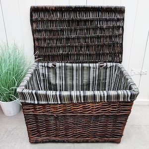 May include: A large, open wicker storage basket with a hinged lid. The basket is a dark brown color and has a striped fabric lining in shades of gray, black, and white. The basket is empty.