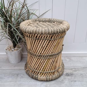 Puede incluir: Un taburete redondo tejido con un marco de bambú natural y una parte superior de cuerda gruesa. El taburete tiene un color marrón natural y está tejido con una cuerda de color marrón más oscuro.