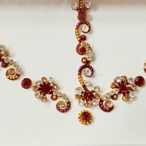 May include: A decorative headpiece or jewelry piece featuring a symmetrical design. The piece is adorned with red and clear gemstones, arranged in floral and circular patterns. Gold-colored accents add contrast against the white background.