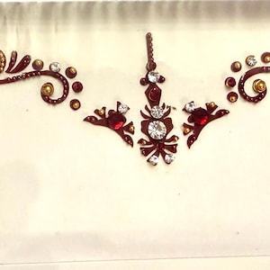 May include: An ornate, decorative forehead bindi design. The design features a series of swirling, floral-inspired patterns in a deep red-brown color, accented with clear and gold-colored rhinestones. The bindi is presented on a white background.