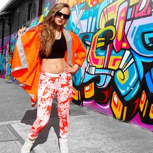 Puede incluir: Una mujer con un chaleco de seguridad naranja brillante sobre un top negro corto y pantalones vaqueros con salpicaduras blancas y rojas. Está de pie frente a una pared con grafitis coloridos.