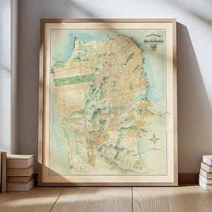 Könnte beinhalten: Eine gerahmte Vintage-Karte von San Francisco, Kalifornien, in Beige-, Grün- und Blautönen. Die Karte mit dem Titel "Chevalier, San Francisco" wird auf einem Holzboden ausgestellt, mit Büchern und einer Pflanze in der Nähe.