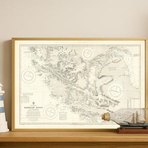May include: A framed map of the Magellan Strait in a gold frame. A decorative lighthouse with blue and white stripes and a ship in a bottle are on a wooden surface. The map is detailed with compass roses.