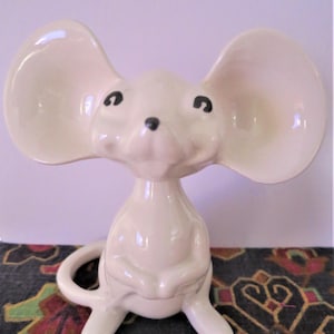 May include: A cream-coloured ceramic mouse figurine in a seated pose. The mouse has large, rounded ears, black eyes, and a small black nose. Its paws are clasped, and it has a curled tail. The figurine is on a patterned surface.
