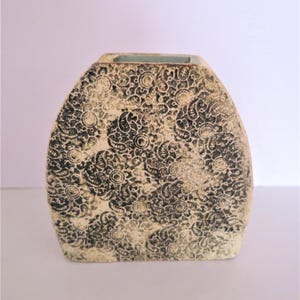 Può includere: Un vaso in ceramica piatto e arrotondato con una superficie testurizzata color crema, caratterizzato da un motivo nero ripetitivo composto da elementi circolari e forme simili al paisley. Il vaso presenta un'apertura rettangolare.
