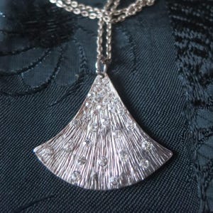以下が含まれることがあります： 扇形のペンダントが付いたシルバーカラーのネックレス。ペンダントは縦線でテクスチャ加工されており、多数の透明な丸いクリスタルで飾られています。ネックレスチェーンもシルバーカラーです。