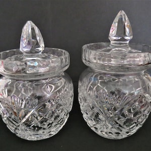 Könnte beinhalten: Zwei klare Glasgefäße mit passenden Deckeln, die jeweils ein detailliertes Diamant- und Fächerschliffmuster aufweisen. Die Gefäße haben einen abgerundeten Körper und einen spitzen Knauf auf jedem Deckel mit reflektierender Oberfläche und Gravuren.