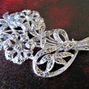 May include: Silver-toned marcasite brooch in the shape of a floral bouquet. The brooch features detailed floral and leaf designs, with a ribbon and bow accent. The marcasite stones add sparkle to the piece.