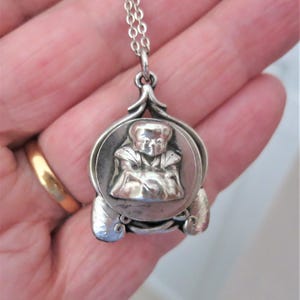 May include: A silver pendant necklace with a detailed, seated figure in a circular frame. The pendant has decorative elements and hangs from a delicate chain. The design is vintage and ornate.