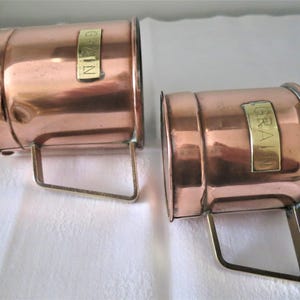 Könnte beinhalten: Zwei Messbecher aus Kupfer mit Messinggriffen und Etiketten mit der Aufschrift "GRAIN". Die Becher haben eine zylindrische Form und stehen auf einer weißen Oberfläche. Das Kupfer hat einen warmen, rötlich-braunen Farbton.