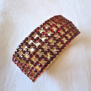 May include: A wide cuff bracelet featuring a gold-tone metal base with a lattice design. The surface is densely encrusted with numerous small, round, deep red faceted rhinestones arranged in a repeating pattern across the entire exterior.