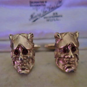 Può includere: Un paio di gemelli dorati, ciascuno con un design dettagliato a teschio e occhi di pietre preziose rosse. I gemelli sono ambientati su uno sfondo morbido e sfocato, suggerendo un'estetica vintage o antica.