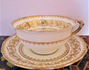 Puede incluir: Juego de taza y platillo de té color crema con detalles florales dorados. La taza tiene un borde festoneado y un asa dorada. El platillo también presenta patrones florales dorados y un borde festoneado. El juego está sobre una superficie estampada.