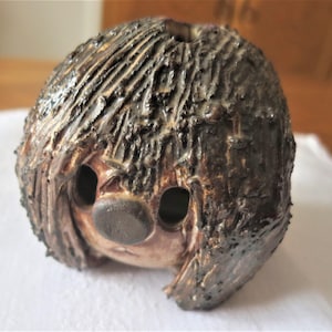 Peut inclure: Une sculpture en céramique faite à la main représentant un visage avec une surface texturée brun foncé ressemblant à des cheveux. Le visage a deux trous pour les yeux et un nez arrondi. La sculpture est une œuvre d'art unique.