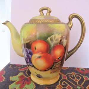 Könnte beinhalten: Vintage Keramik-Teekanne mit handgemaltem Fruchtdesign. Die Teekanne hat einen gelben Sockel, Ausguss und Griff mit goldenen Akzenten. Der Korpus ist mit Pfirsichen und Trauben verziert. Der Deckel hat einen goldenen Griff.
