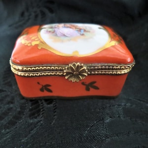 Könnte beinhalten: Kleine, rechteckige, orangefarbene Schmuckschatulle mit Scharnierdeckel. Der Deckel zeigt eine gemalte Szene in einem weißen Oval, umrahmt von goldenen Akzenten. Die Schatulle hat goldene Verzierungen und einen floralen Verschluss. Schwarze Blattmotive zieren die Seiten.