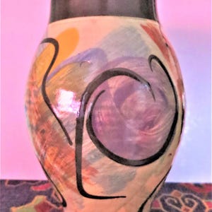May include: A hand-painted ceramic vase with a black rim and abstract design. The vase features a cream-coloured base with colourful brushstrokes of orange, purple, blue, and red, accented by black swirling lines. The vase is approximately 20 cm tall.