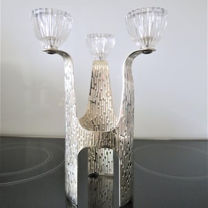 May include: A silver candelabra with three clear glass candle holders. The candelabra features a textured, vertical line design and a modern shape. The candle holders are bowl-shaped with a ribbed texture.