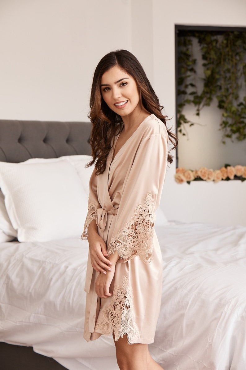 May include: A light beige satin robe with delicate lace trim on the sleeves and hem. The robe has a V-neck and a tie belt.