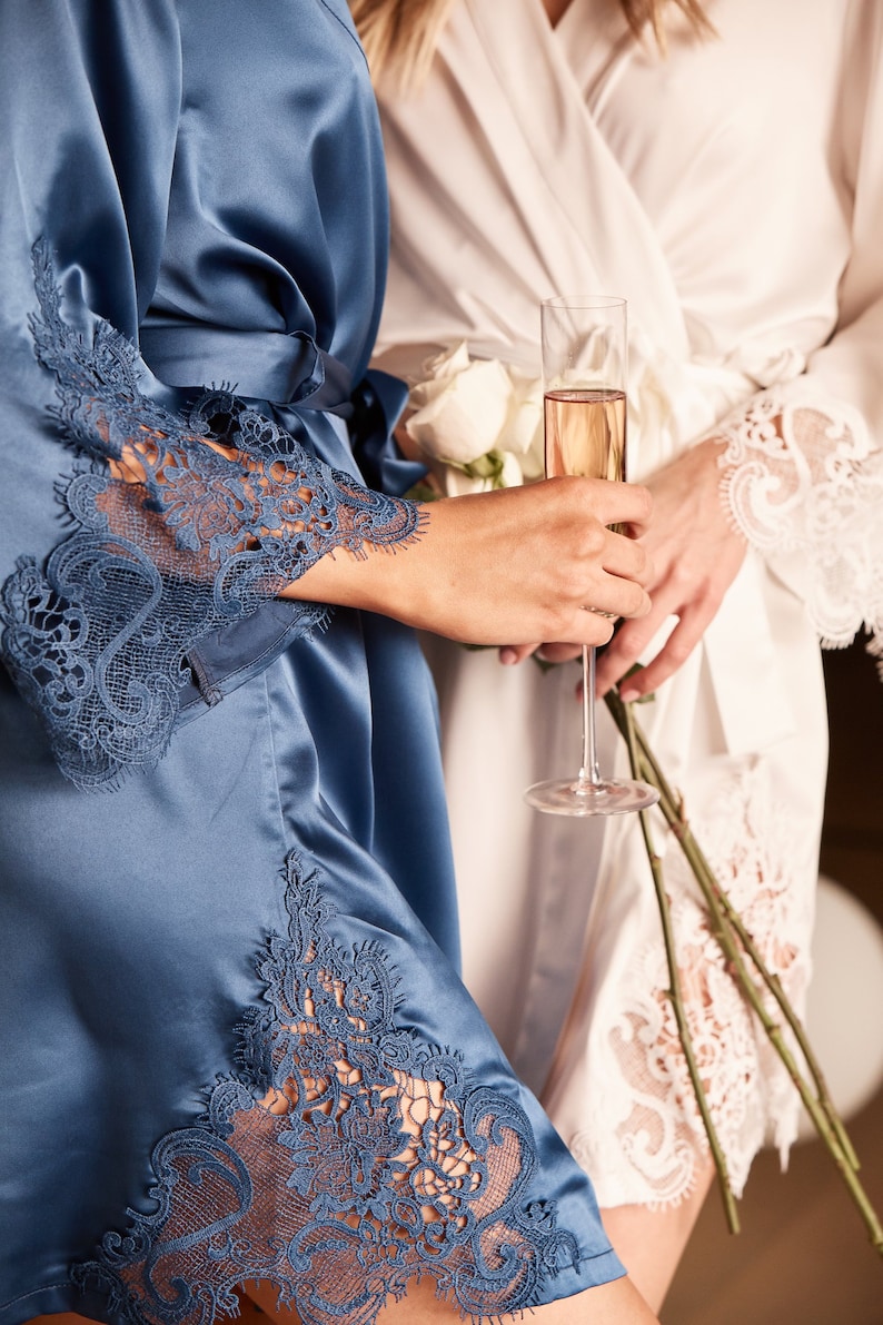May include: Two women wearing satin robes with lace trim. One robe is blue and the other is white. The woman in the blue robe is holding a glass of champagne.