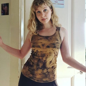 May include: A woman wearing a brown and gold tie-dye tank top with black leggings. The tank top has a relaxed fit and a scoop neck. The woman is standing in front of a white door with a quote from Rumi on it.