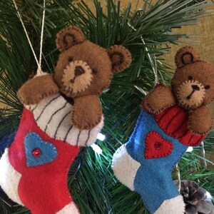 May include: Two felt Christmas stocking ornaments with brown teddy bears peeking out of the top. The stocking on the left is red with a blue heart and the stocking on the right is blue with a red heart.