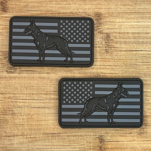 Pode incluir: Duas etiquetas retangulares com a silhueta de um cão sobre um desenho de bandeira americana cinza e preta. O cão está em pé, e a bandeira tem estrelas e listras. As etiquetas estão em uma superfície de madeira.