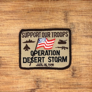 May include: A rectangular patch with the text "SUPPORT OUR TROOPS" and "OPERATION DESERT STORM JAN. 16, 1991". The patch features an American flag, a jet, a tank, and a rifle. The patch has a tan background with a brown border.